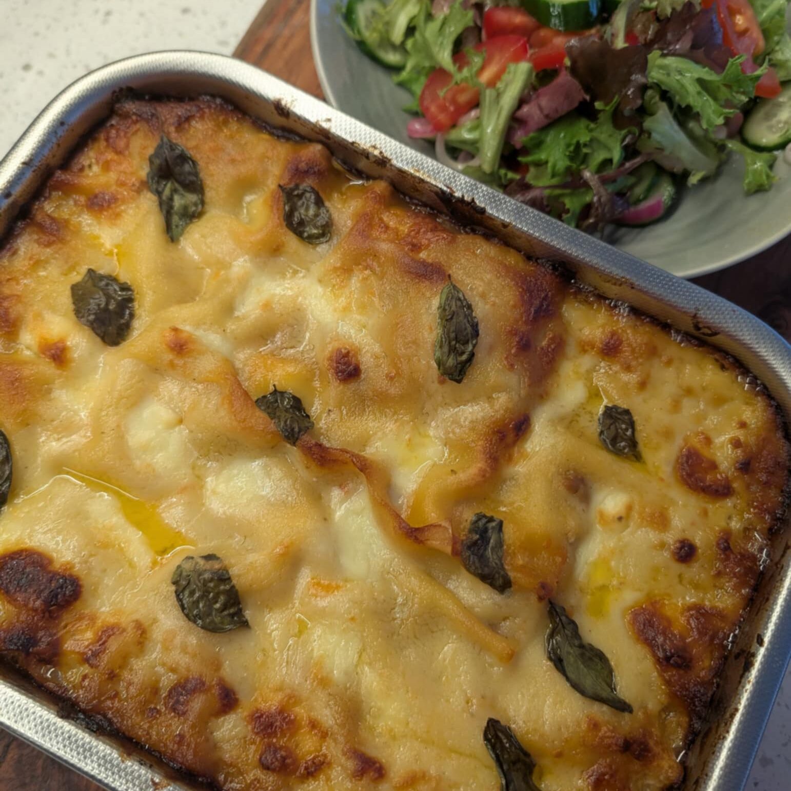 photo of a golden brown cooked lasagne in an oven dish with salad in the background