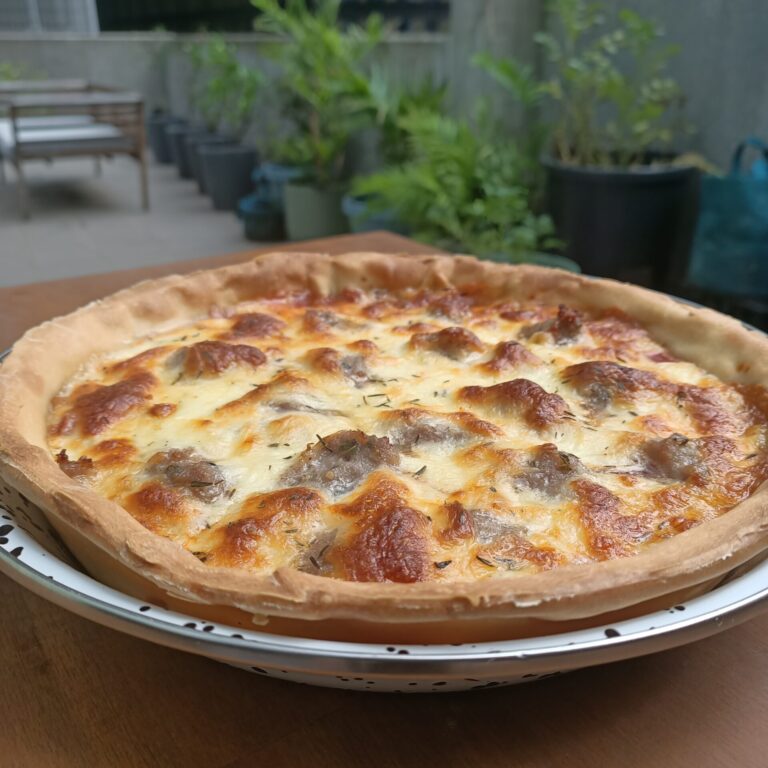 photo of a pizza in a metal tray on a wooden bench plants in background